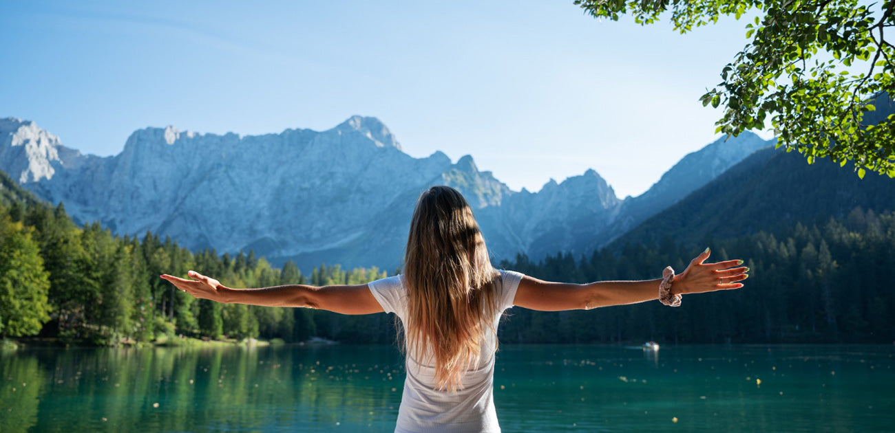 A joyful woman embraces the tranquility of nature, positioned before a lake with outstretched arms.